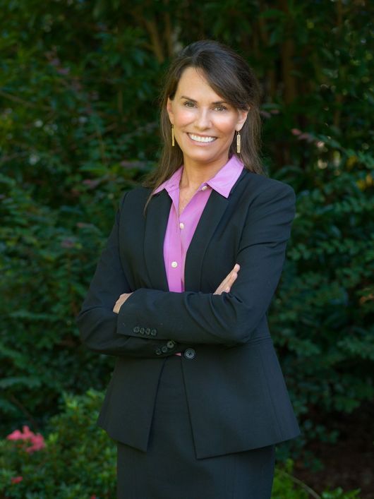 A person in a black blazer and pink collared shirt smiling with arms crossed in front of a green leafy backdrop.
