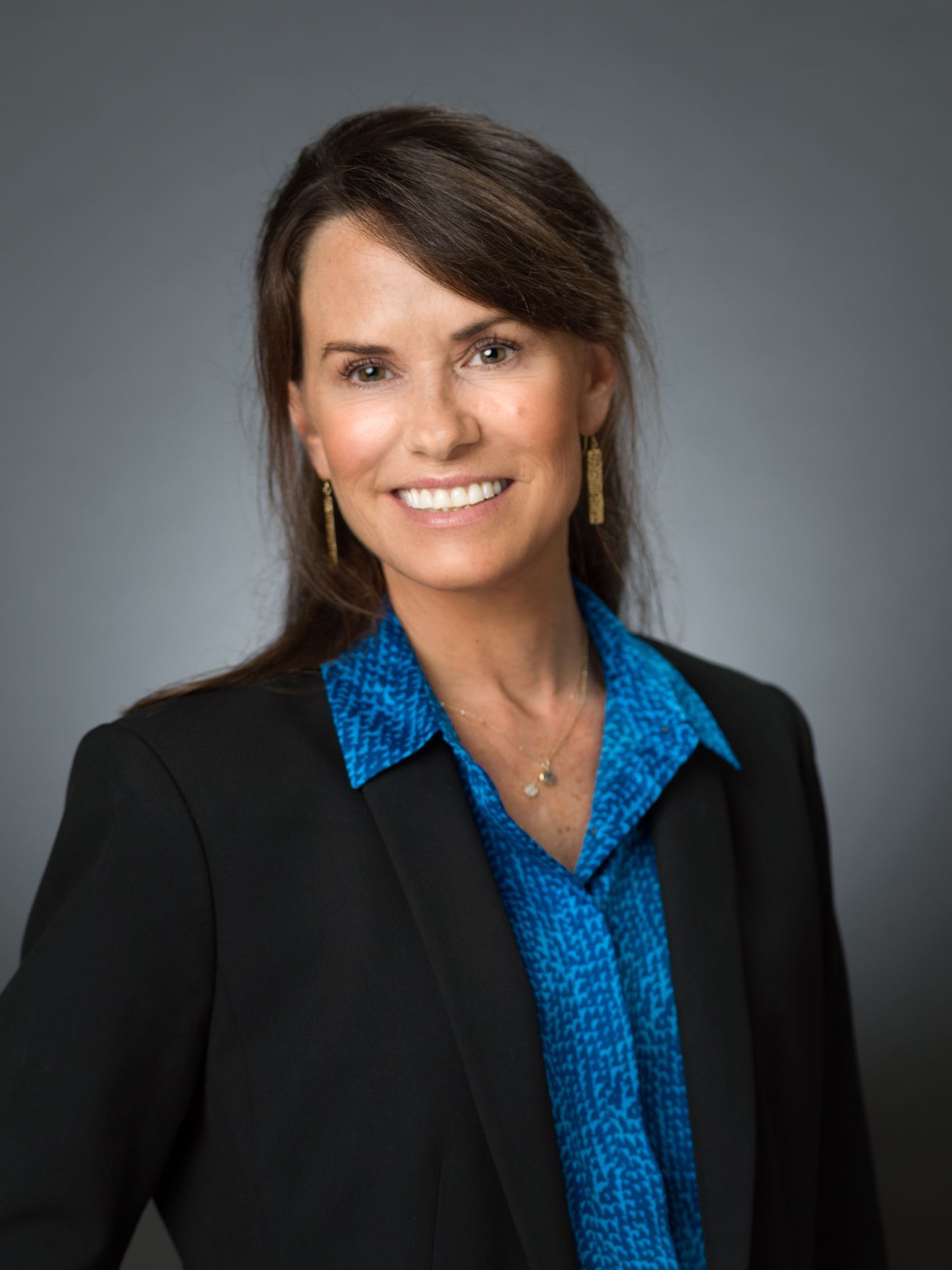 Professional headshot of a person smiling, wearing a blue patterned shirt under a black blazer against a gray background.