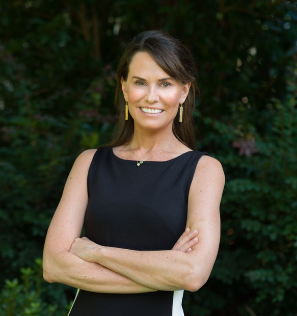 A smiling woman with dark hair and gold earrings wears a black sleeveless dress with her arms crossed outdoors.