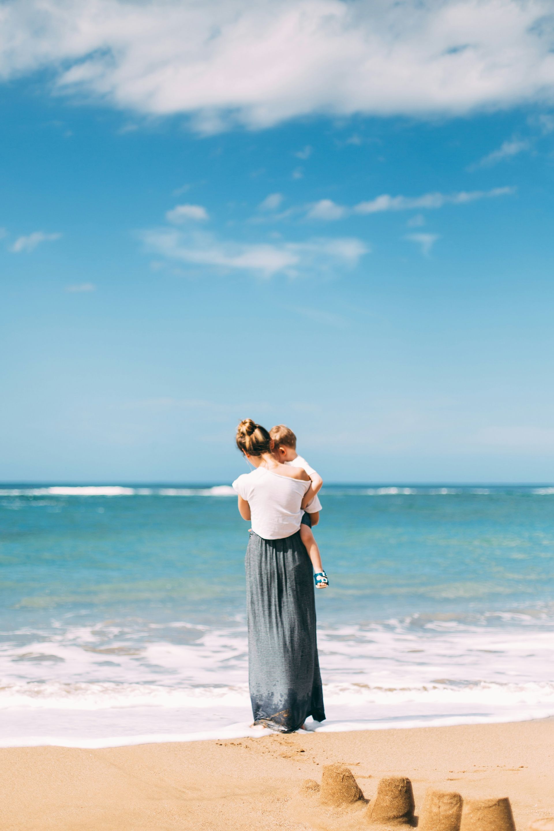 A person carrying a child stands on a sandy beach, looking out at the turquoise ocean under a bright blue sky.
