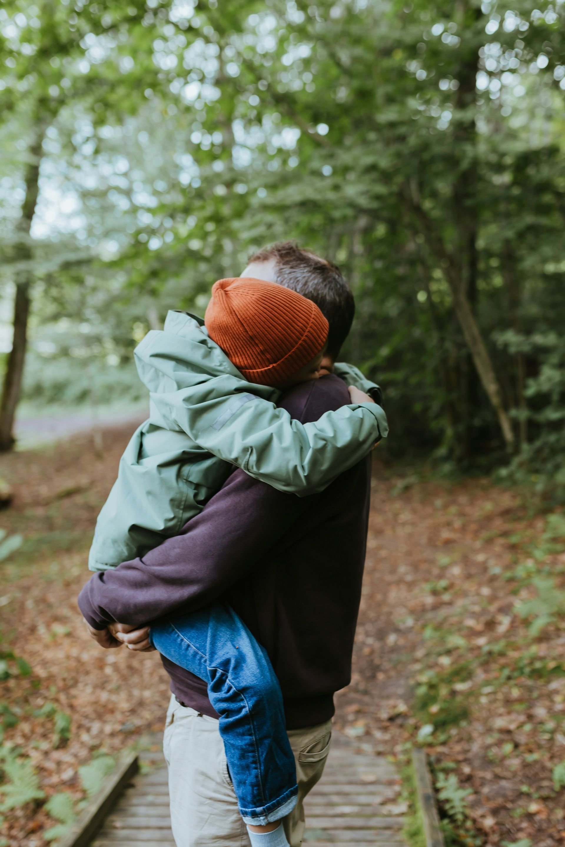 A person holding a child wearing a green coat and orange beanie on a wooden path in a wooded area.