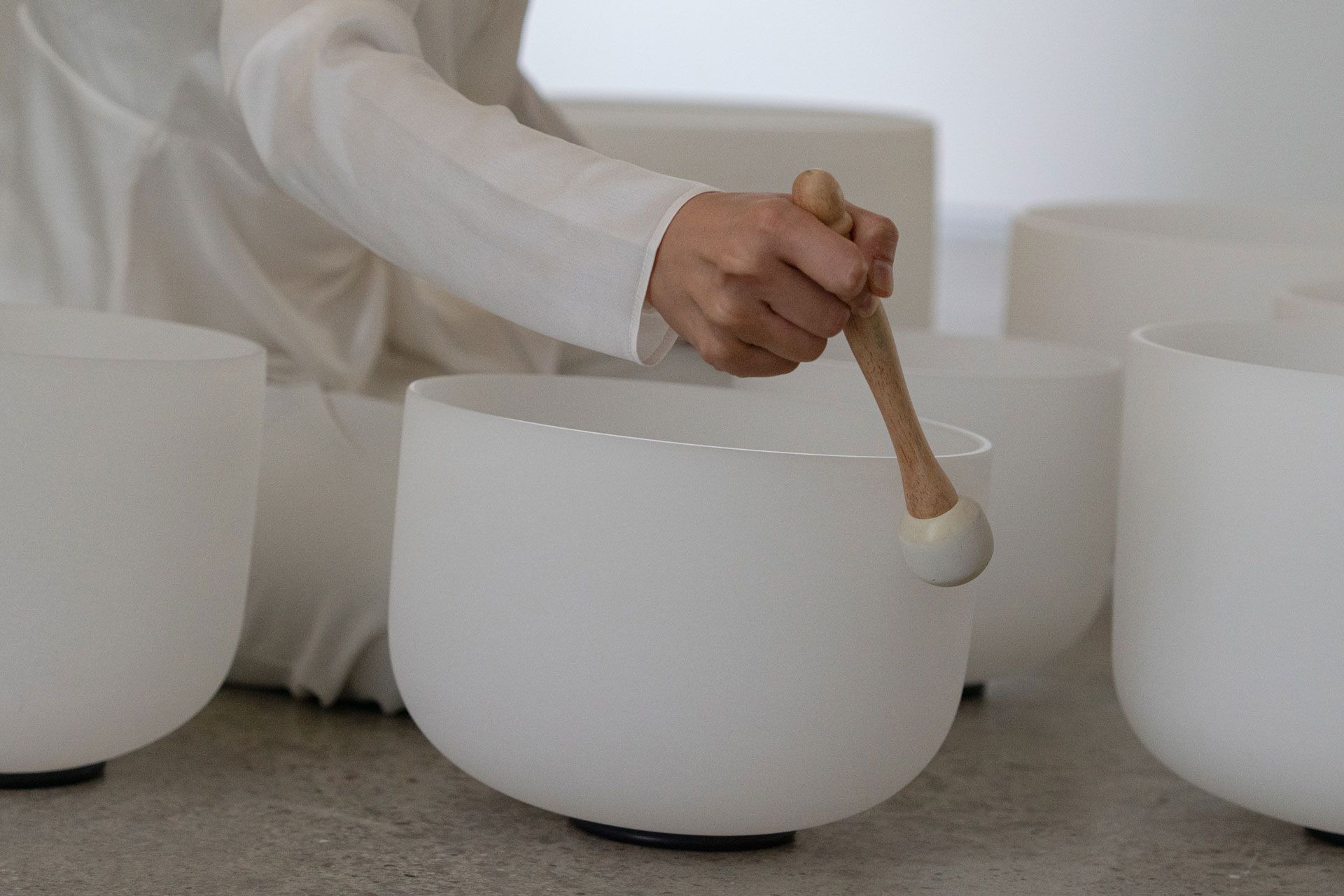 Person in white clothes playing a crystal singing bowl with a mallet. Several bowls are visible.