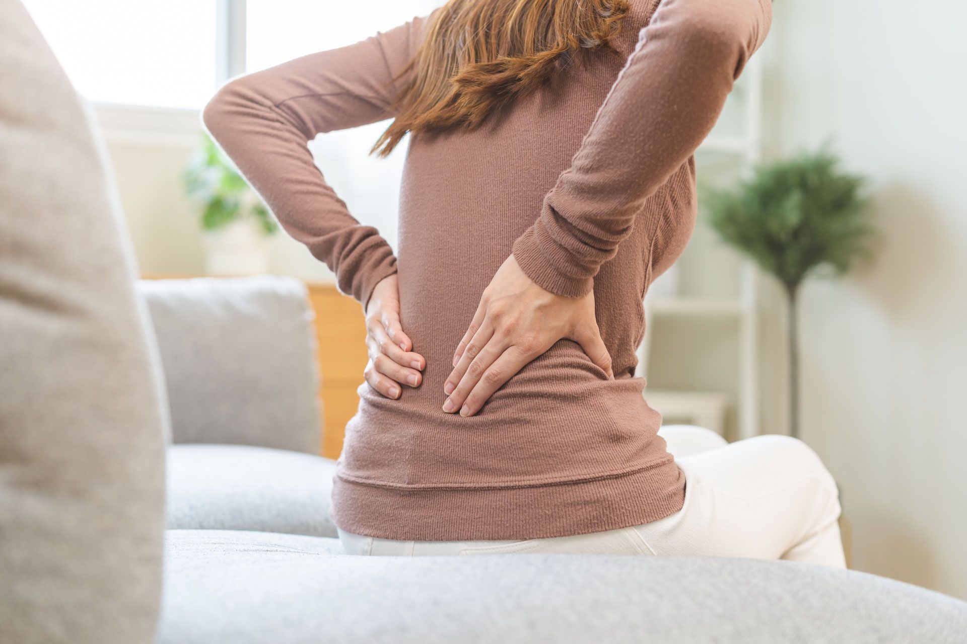 A person sitting on a sofa, leaning forward and holding their lower back with both hands due to discomfort.
