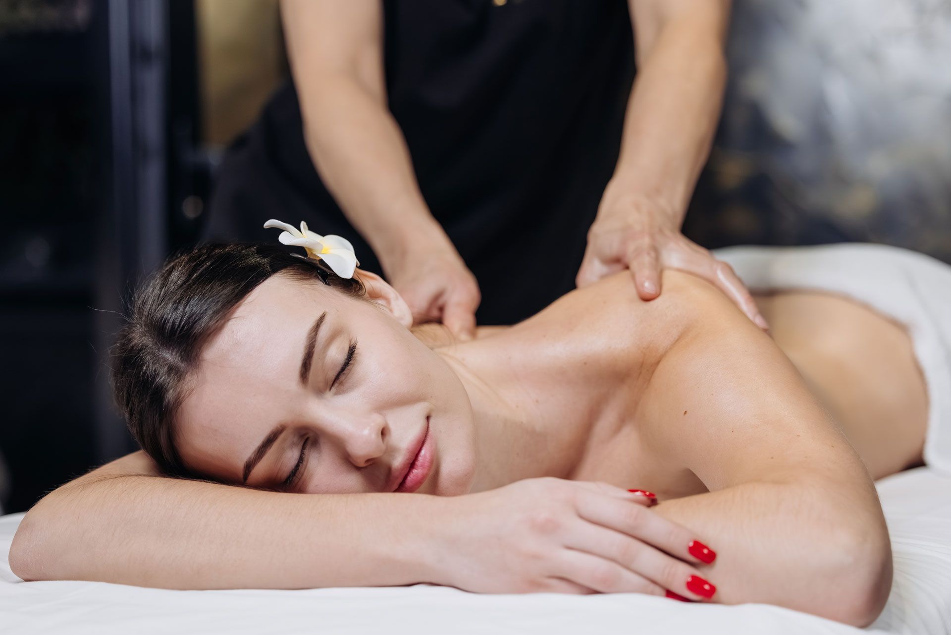 A person receiving a shoulder massage while lying on a spa table with a small white flower in their hair.