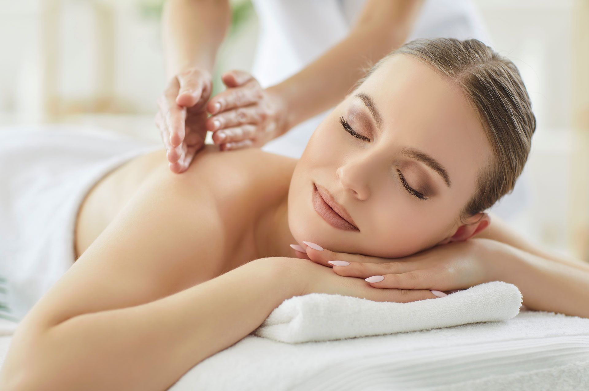 A person with eyes closed lies on a spa table, receiving a back massage with their arms resting on a white towel.