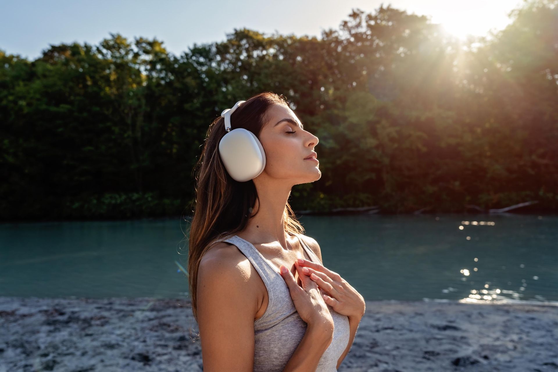 Woman with headphones, eyes closed, hands on chest, by a river, sunlit trees in the background.