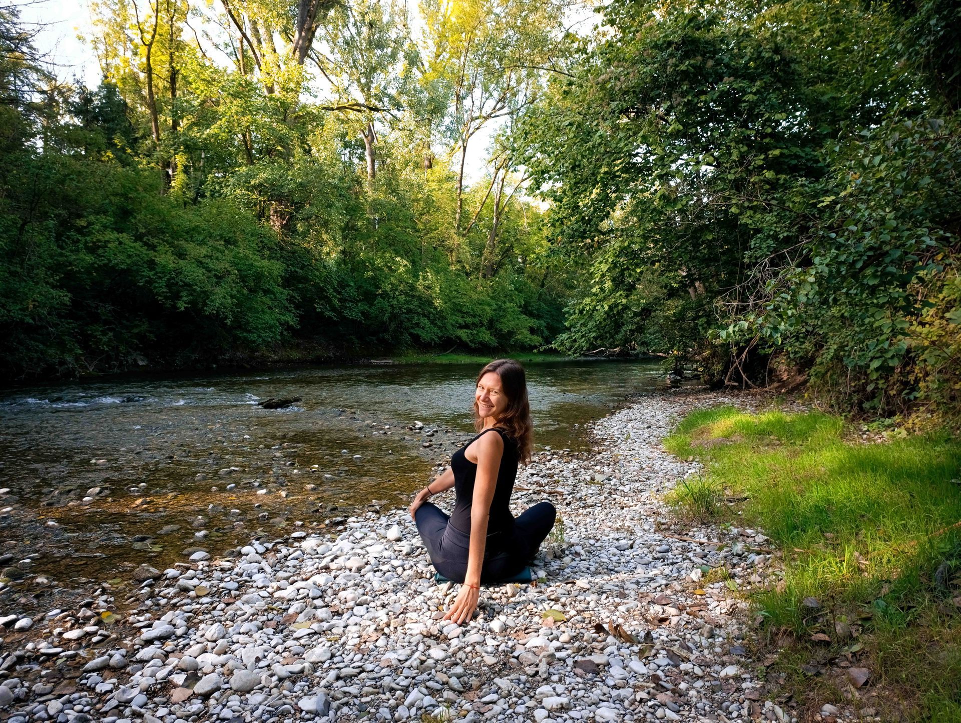 Frau in Yoga-Pose an einem Fluss, umgeben von Bäumen und Grün.