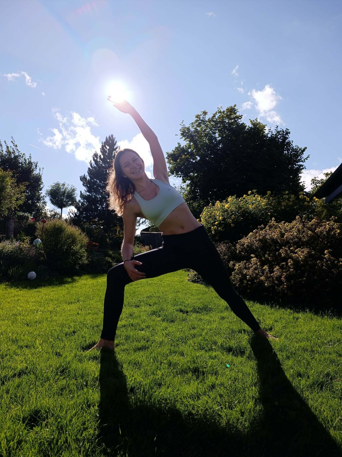 Frau in Yoga-Pose, lächelnd, mit erhobenem Arm zur Sonne. Draußen auf Gras, sonniger Tag.