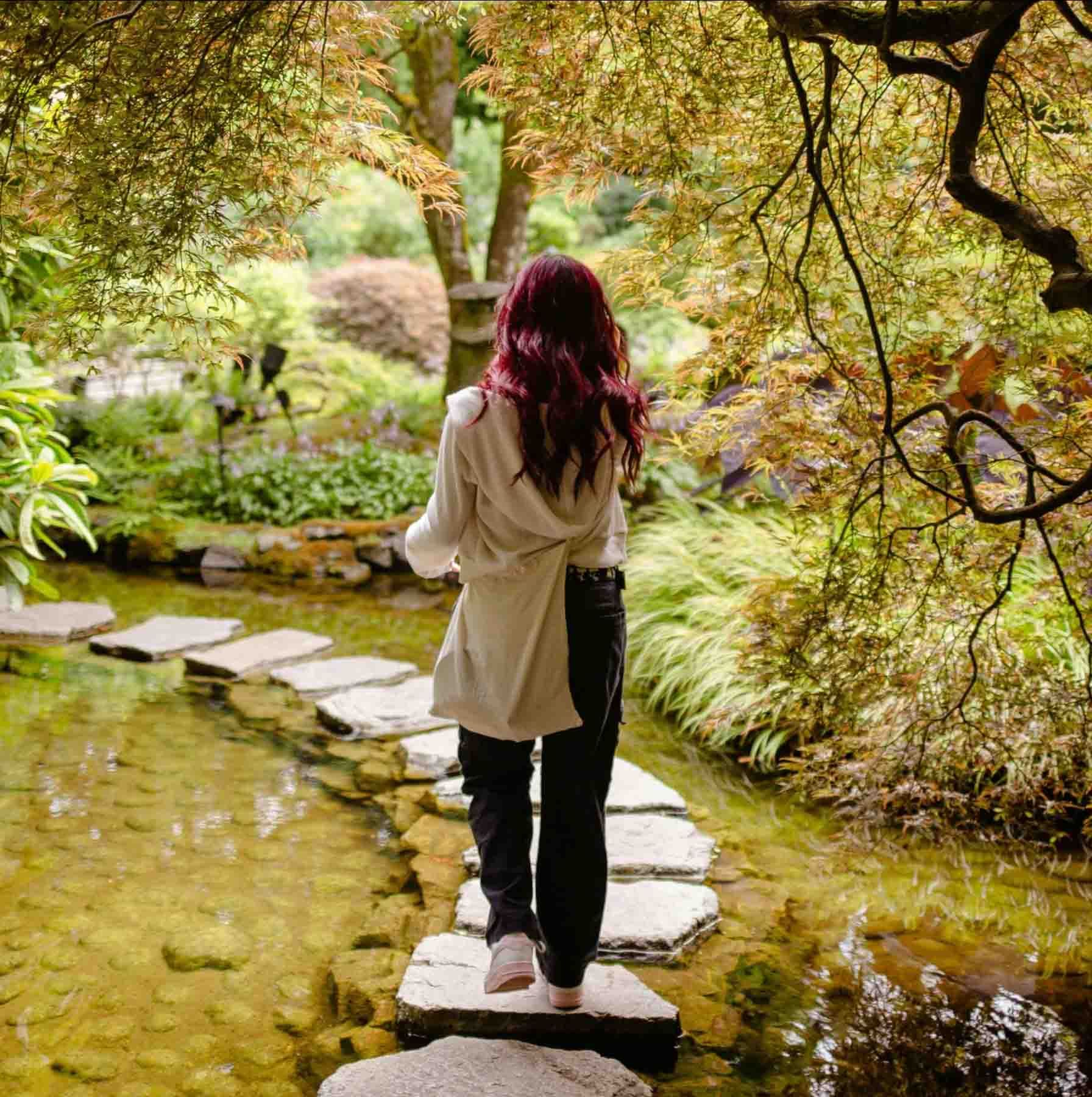 Frau mit roten Haaren geht über Trittsteine ​​in einem japanischen Garten, umgeben von Laub.