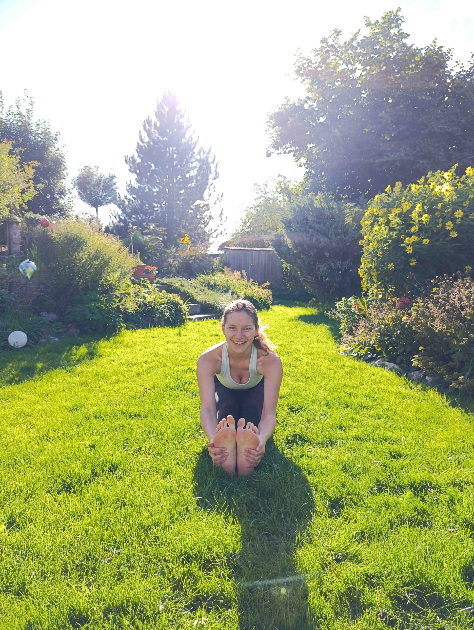 Frau in Yoga-Pose auf Gras, streckt ihre Füße im sonnigen Garten aus.