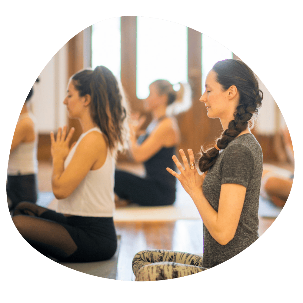 Frauen im Yoga-Kurs, sitzend, die Hände zum Gebet geformt, die Augen geschlossen. Drinnen, Holzboden, Tageslicht.