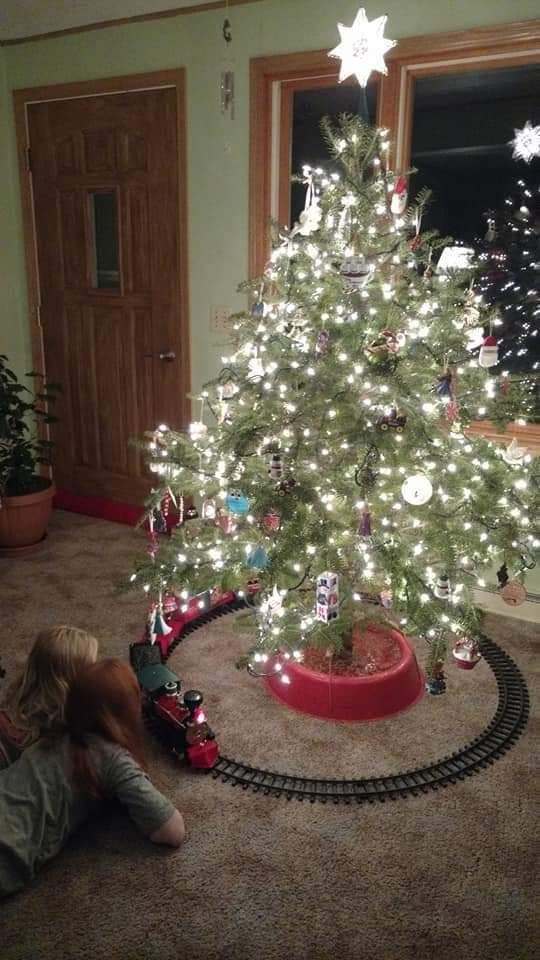A little girl is playing with a train around a christmas tree.