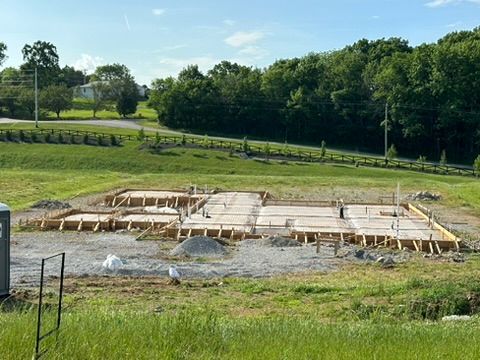 Wooden foundation formwork on a gravel base in a grassy field. Trees and a blue sky in the background.