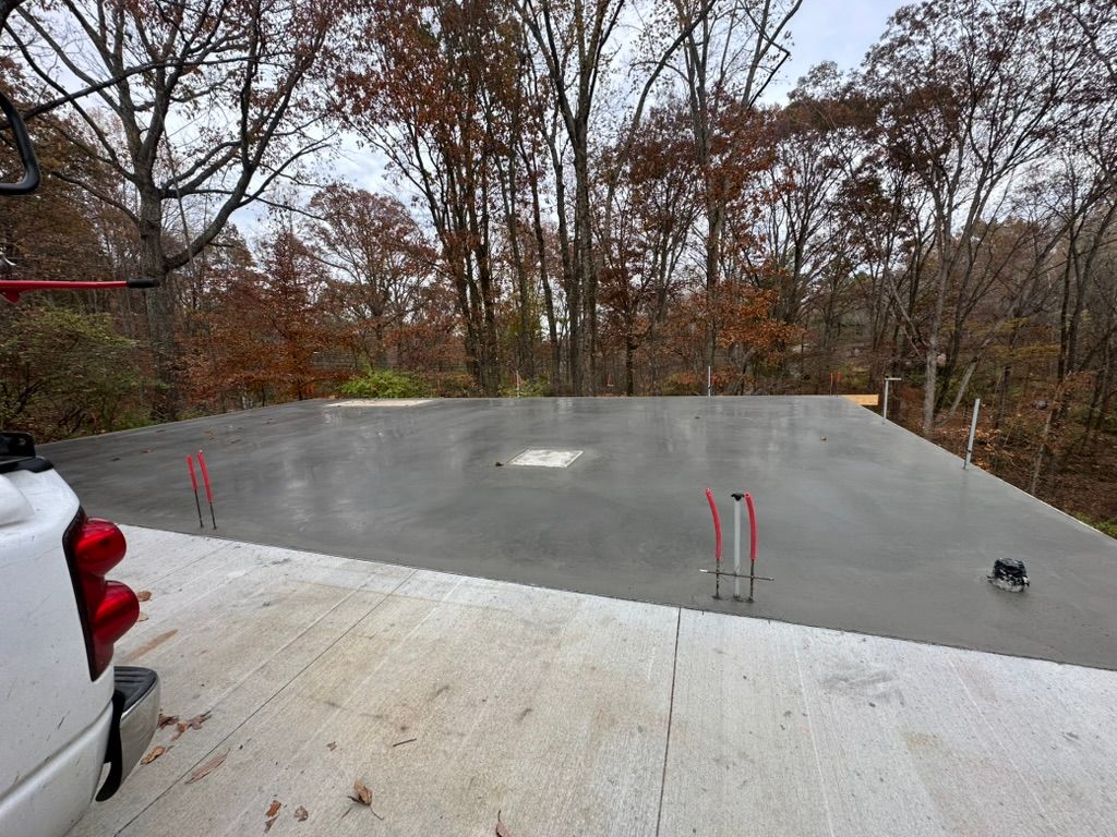 Freshly poured concrete foundation with red pipes, truck bed in foreground, trees in background.