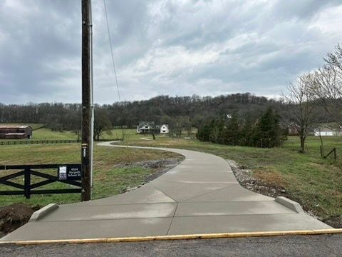 Newly poured concrete driveway curves toward a house in a rural landscape under a cloudy sky.