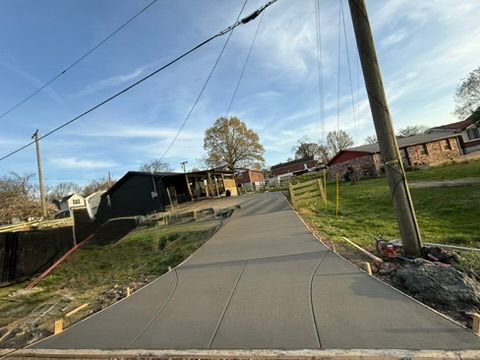 Newly poured concrete pathway on a slight incline, flanked by grass, buildings, and utility poles under a blue sky.