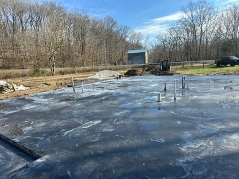 Newly poured concrete foundation with pipes, construction site in the background. Blue sky, bare trees.