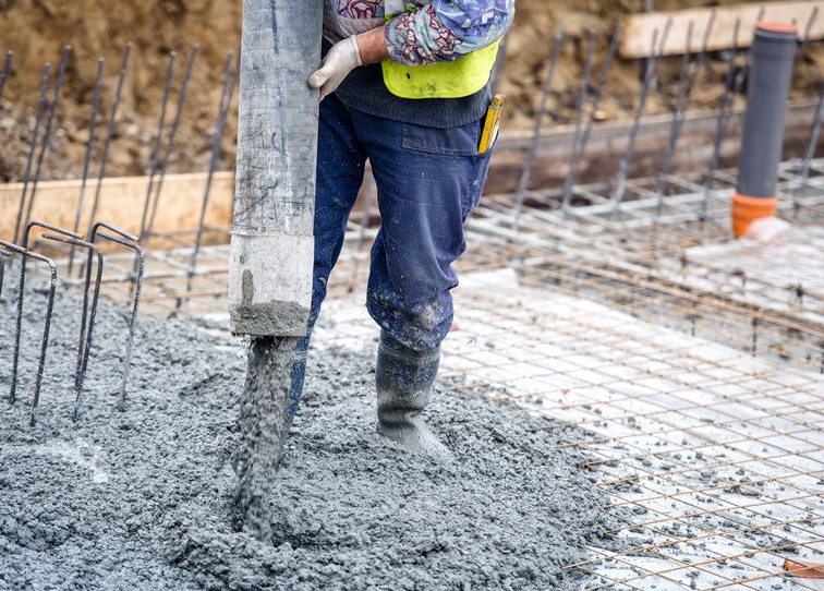 Construction worker pouring concrete at a building site, wearing boots and safety vest.