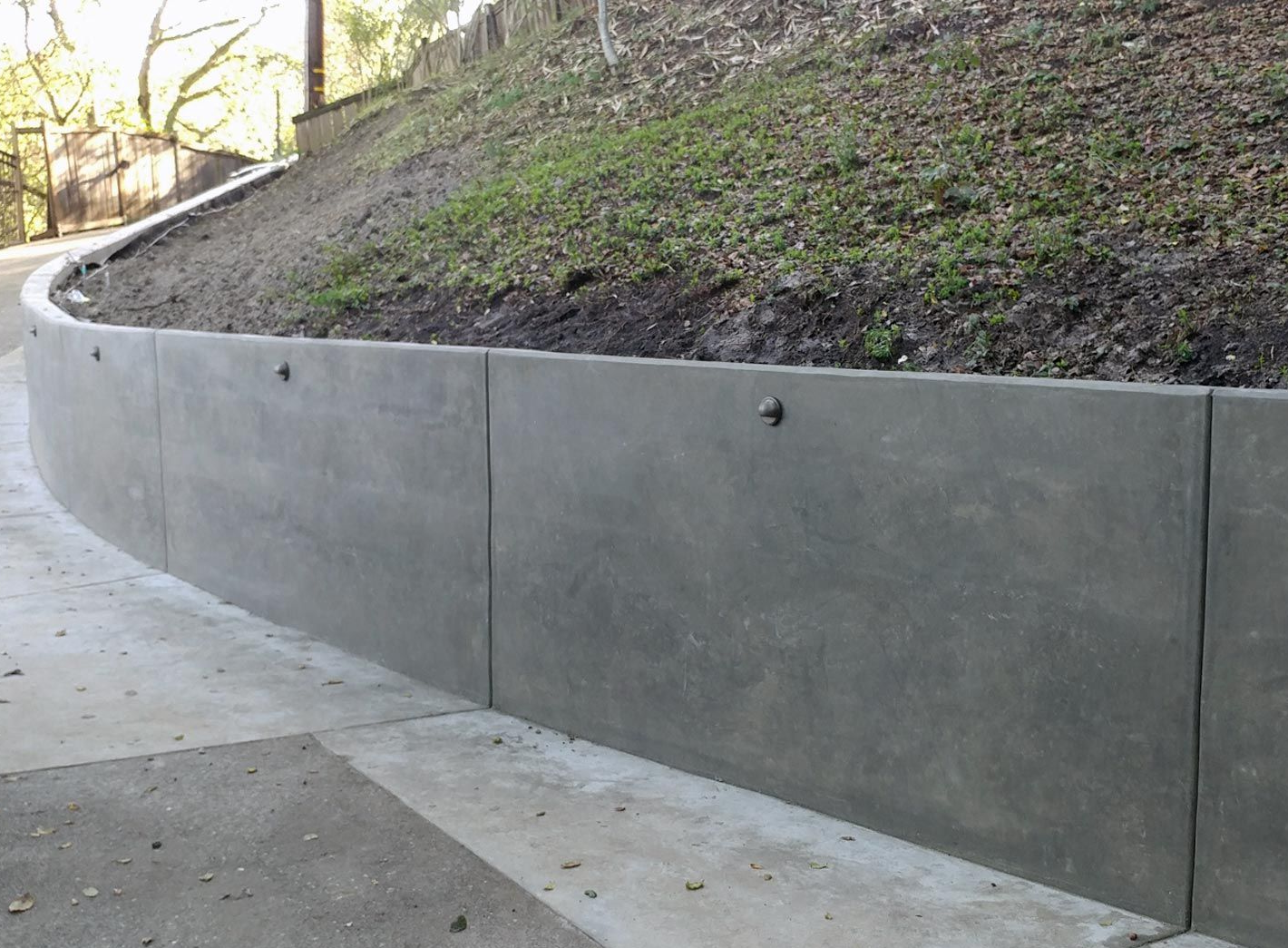 Gray concrete retaining wall alongside a paved road, supporting a grassy hillside.