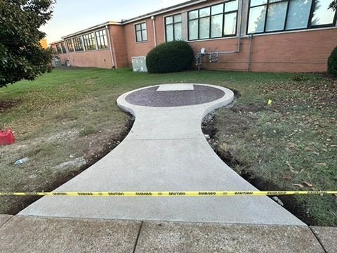 Concrete sidewalk leading to a circular platform with gravel; caution tape across the path; brick building in the background.