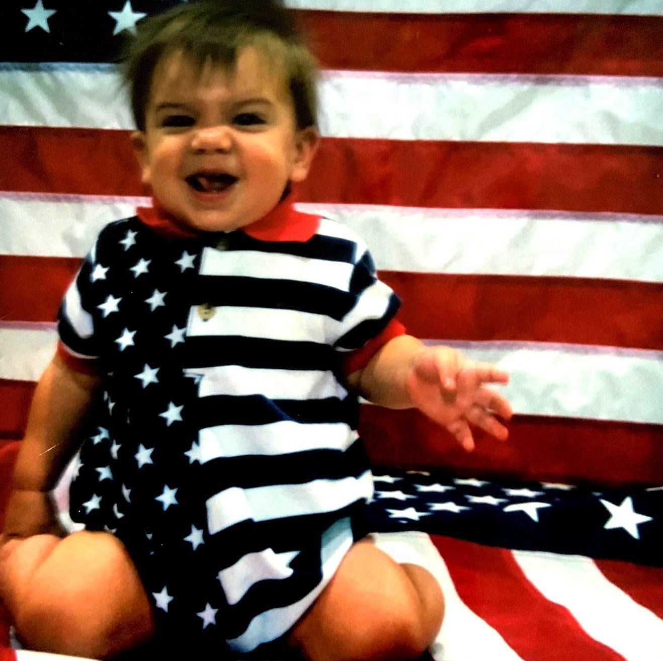 A baby in an american flag outfit is sitting in front of an american flag