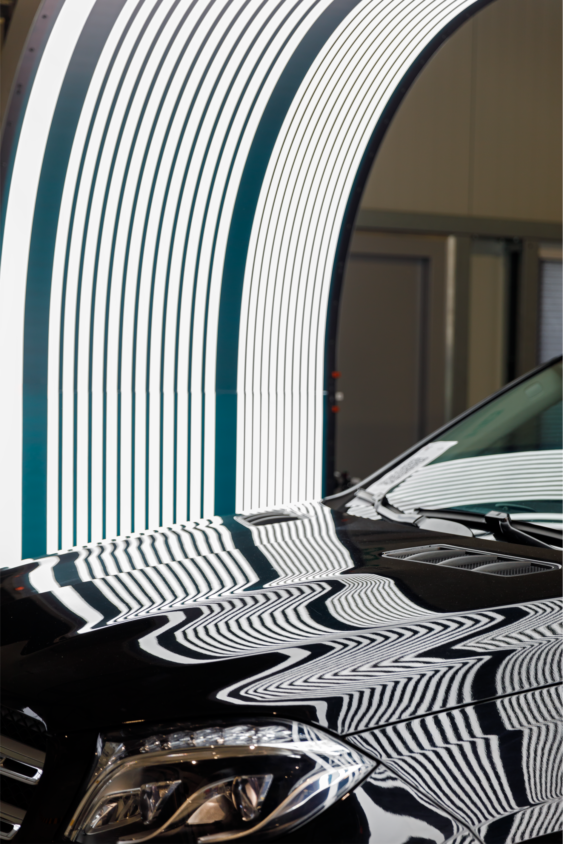 A black luxury car parked under a light tunnel, reflecting the bright white and dark stripe patterns on its glossy hood.