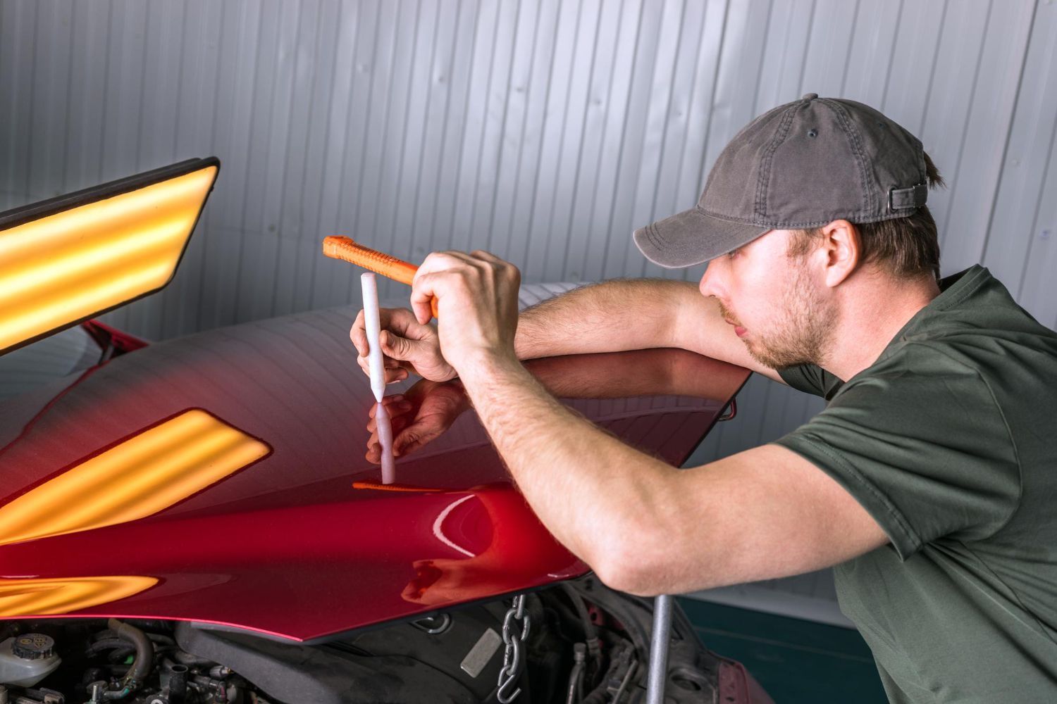 Man in cap using a tool to remove a dent from a red car hood.