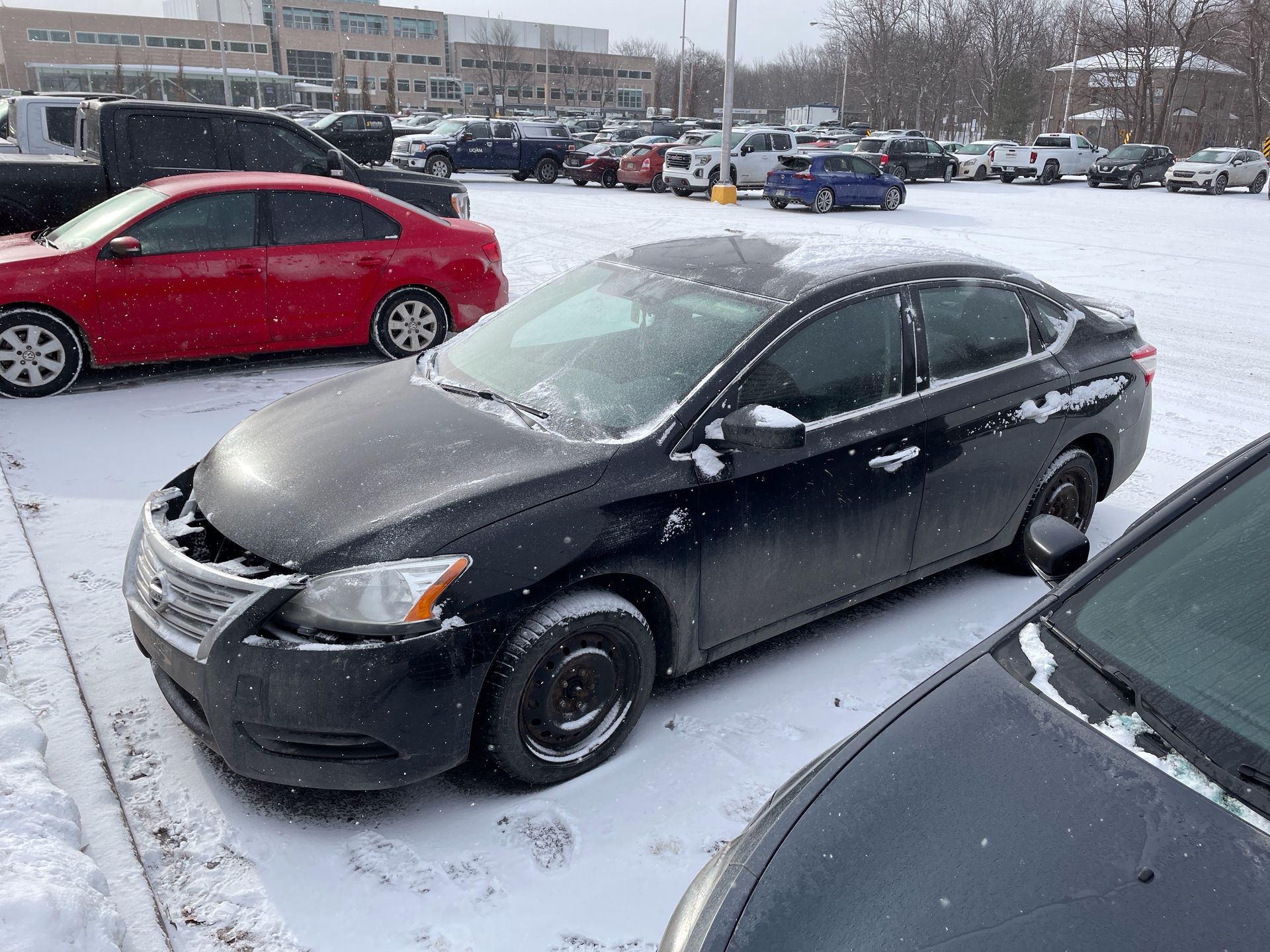 Black sedan in a snowy parking lot with missing front bumper, surrounded by other cars.