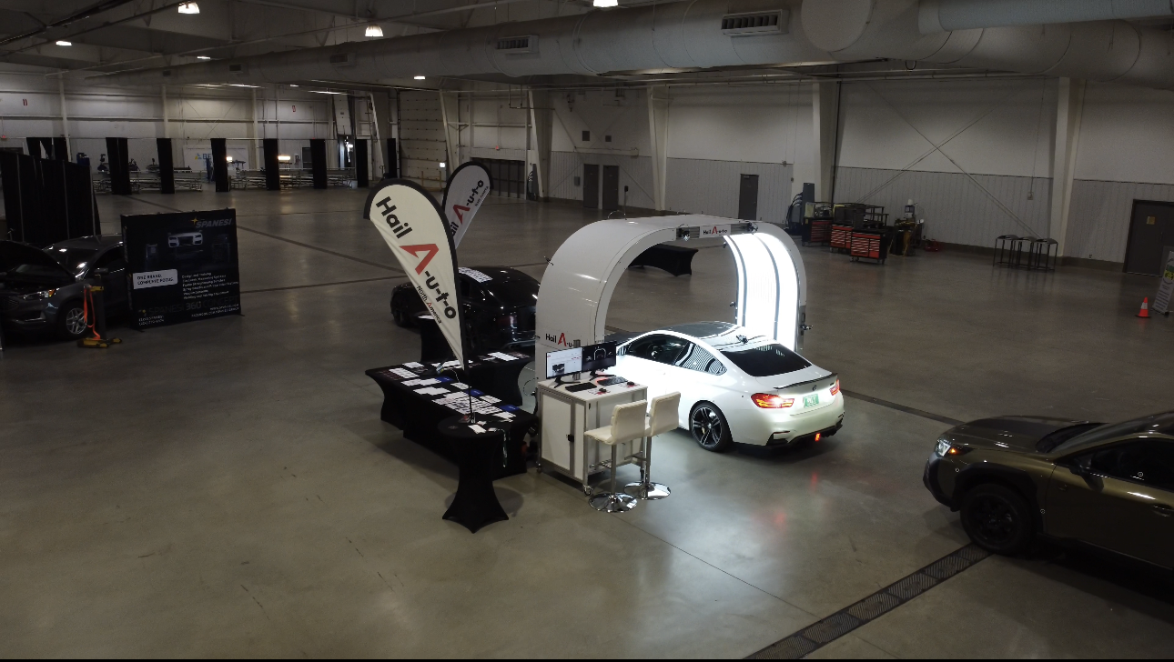 A white car parked under a bright arch, inside a large event space with promotional materials.