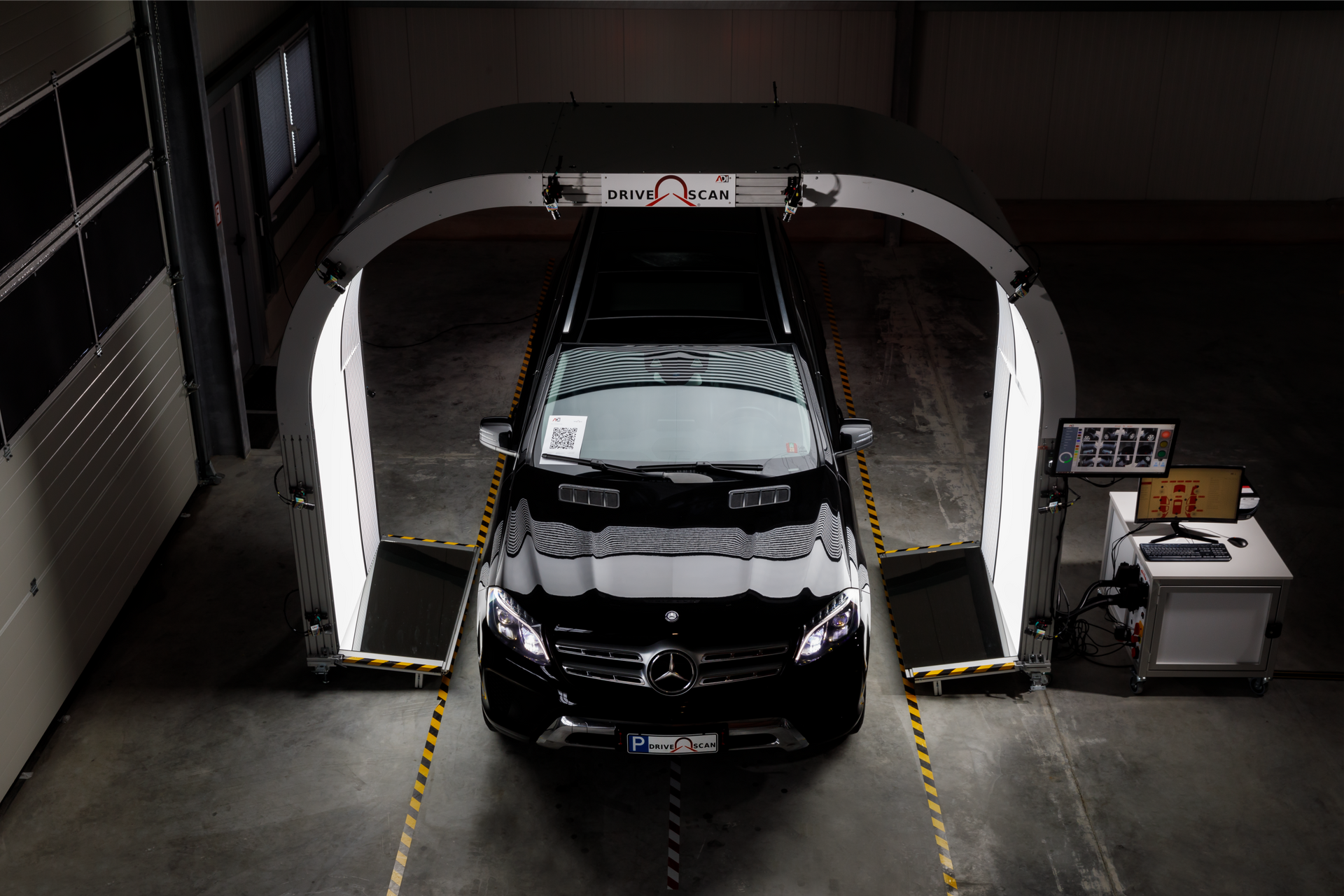 A black Mercedes car sits inside a diagnostic inspection portal with overhead lights and a computer workstation nearby.