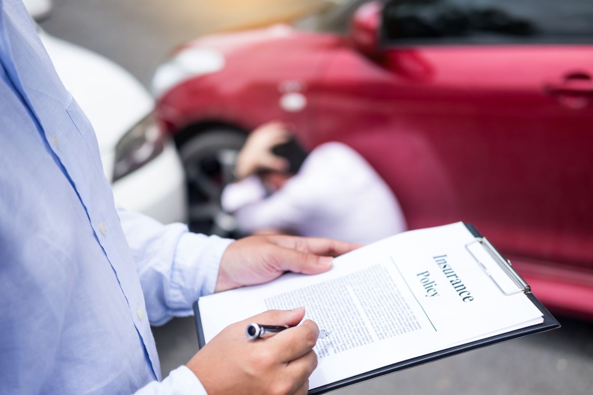 Person holding insurance policy form at a car accident scene, red and white cars damaged.