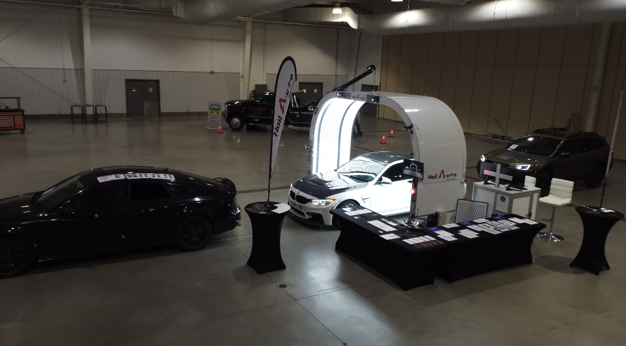 White enclosed car detail booth with car, dark cars, and display tables in a large room.