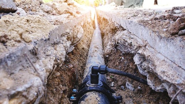 A trench cut through concrete pavement, exposing a dark pipe with a service connection fitting attached.