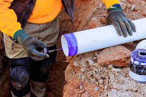 A construction worker in a high-visibility orange jacket uses a dauber to apply purple primer to a white PVC pipe.