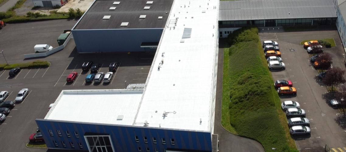 Vue aérienne de bâtiments industriels avec parkings et voitures stationnées. Présence de buissons verts.