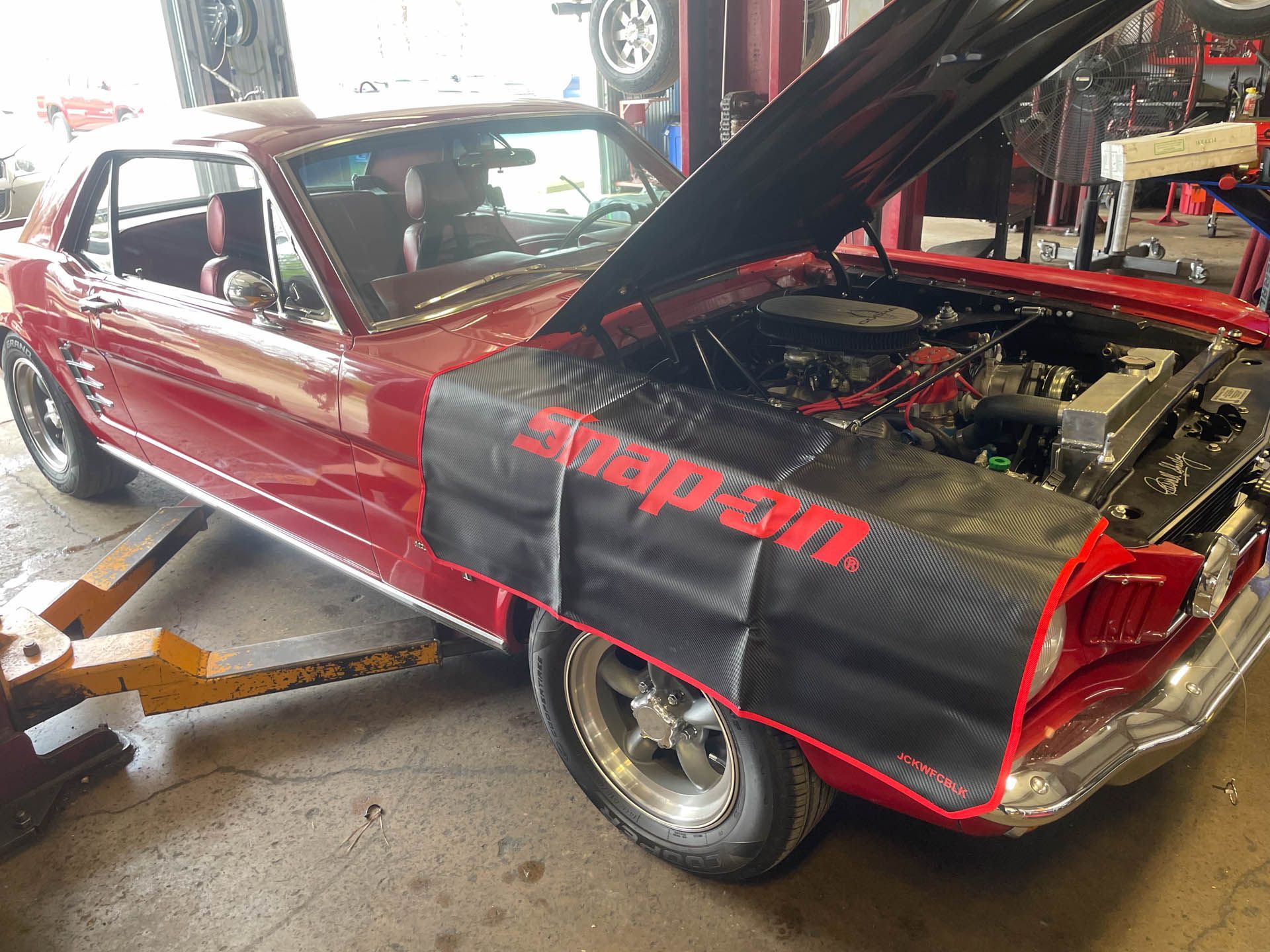 A red car with the hood up is sitting on a lift in a garage.