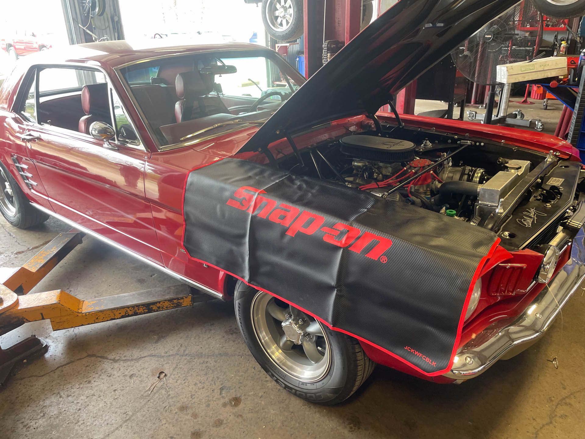 A red car with the hood up is sitting on a lift in a garage.