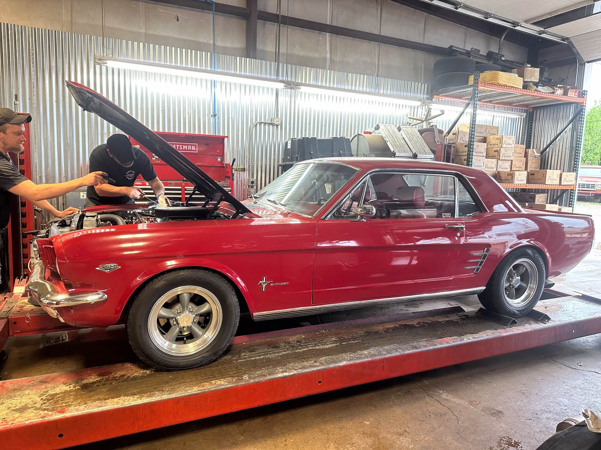Two men are working on a red mustang in a garage.