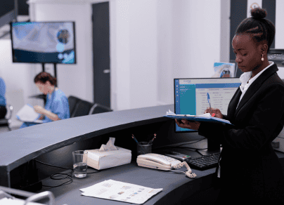 A woman is sitting at a desk in front of a computer holding a clipboard.