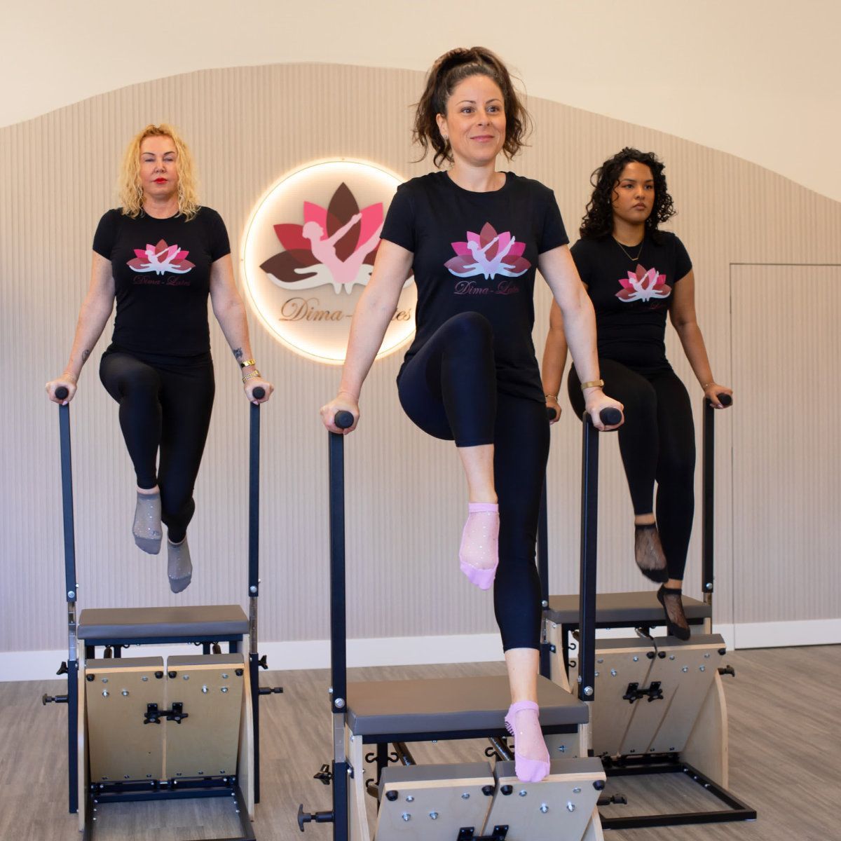 Three women in black workout attire exercising on Pilates chairs. Inside a studio, lotus logo on the wall.