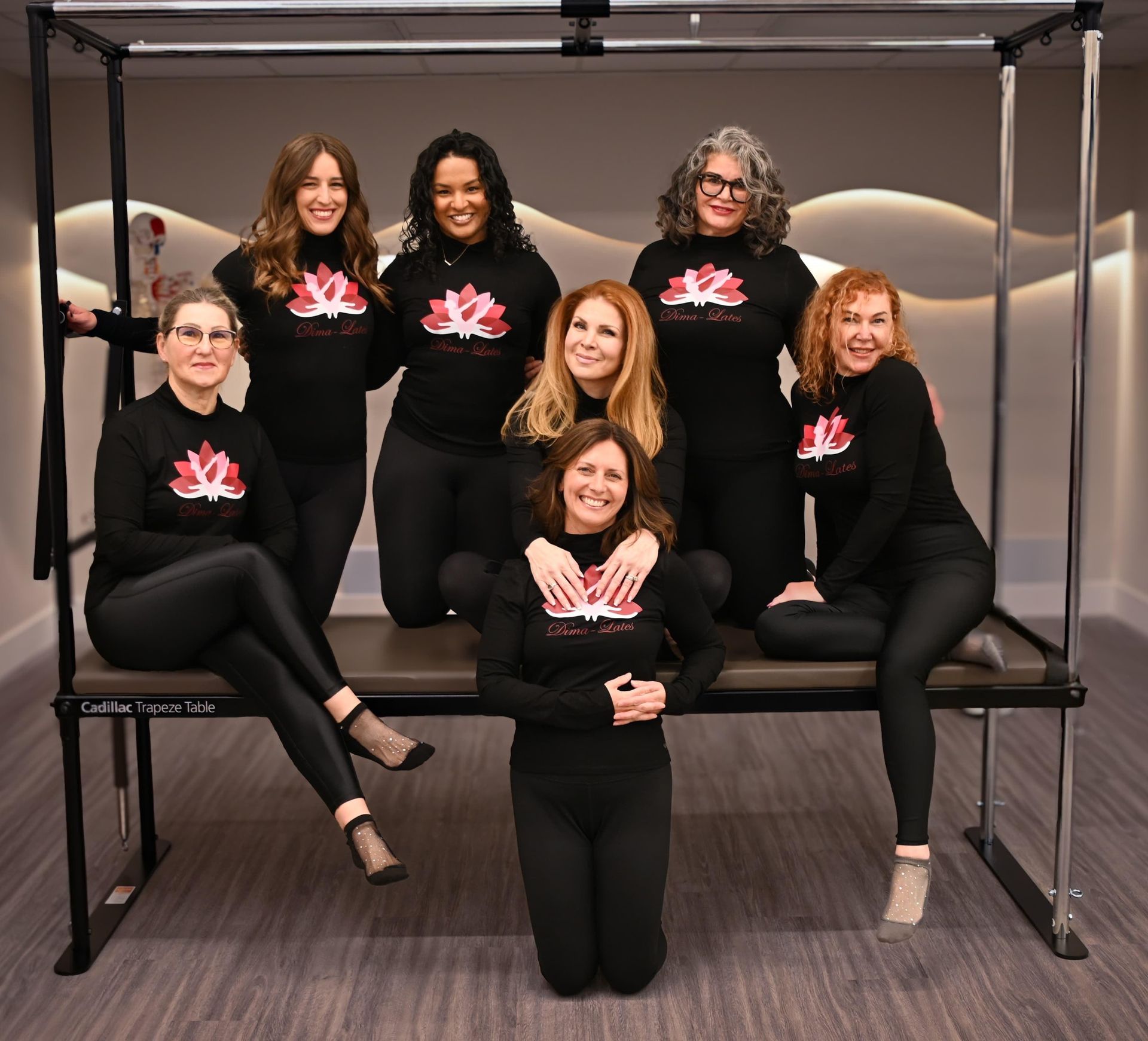 Group of women posing in a pilates studio. They are wearing matching pink t-shirts and black leggings.