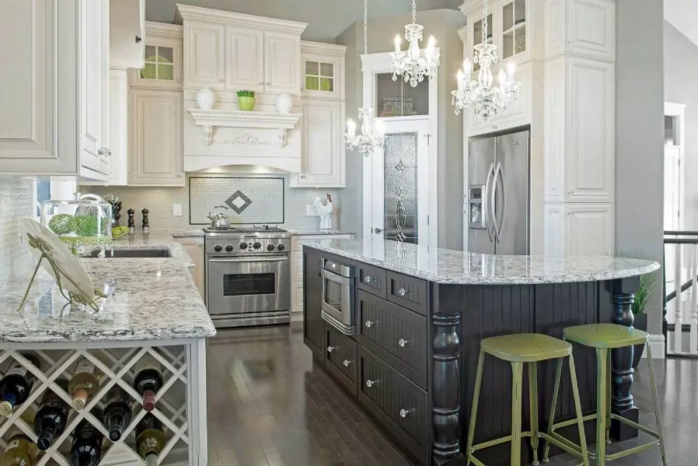 Elegant kitchen with white cabinets, dark island with seating, stainless steel appliances, and two chandeliers.