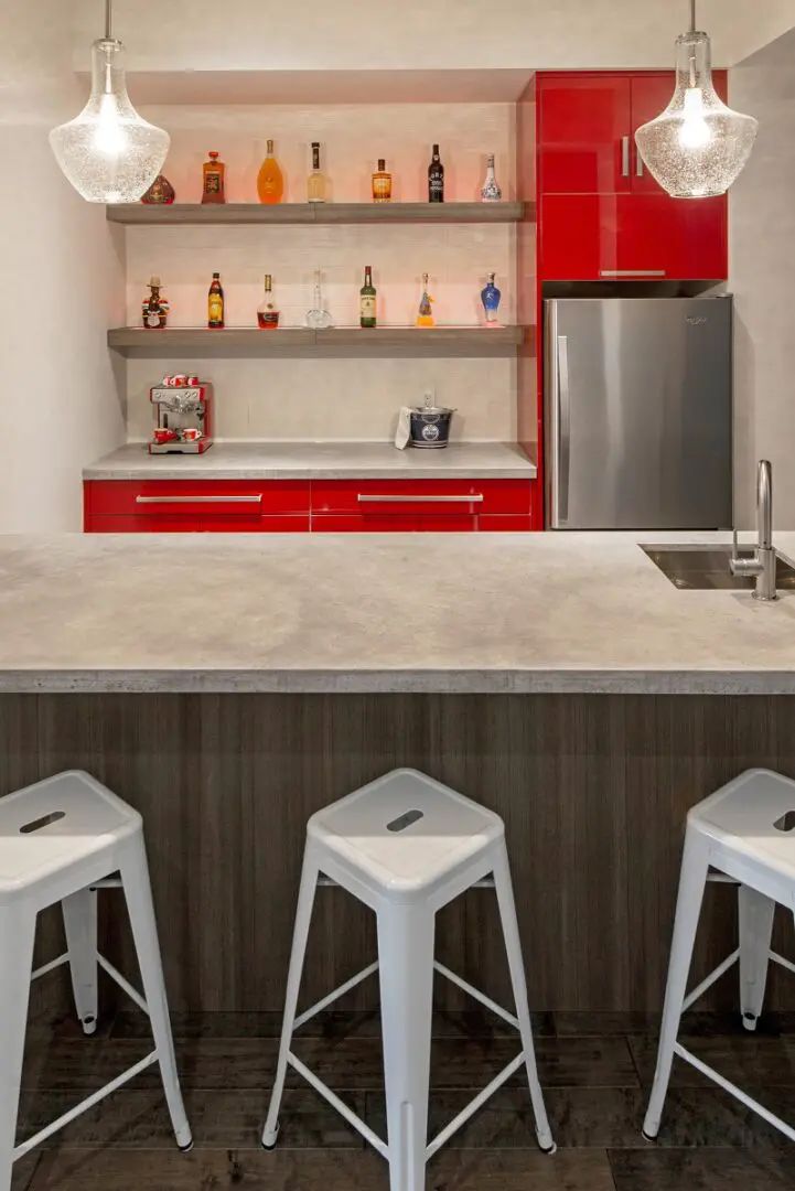 Red and white home bar with liquor bottles on shelves, stainless steel fridge, and white stools.