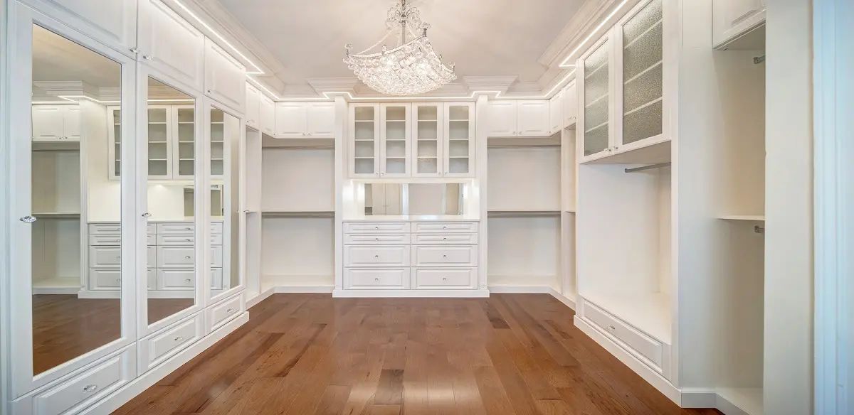 Spacious, white walk-in closet with hardwood floors, a large chandelier, and built-in shelving and cabinets.