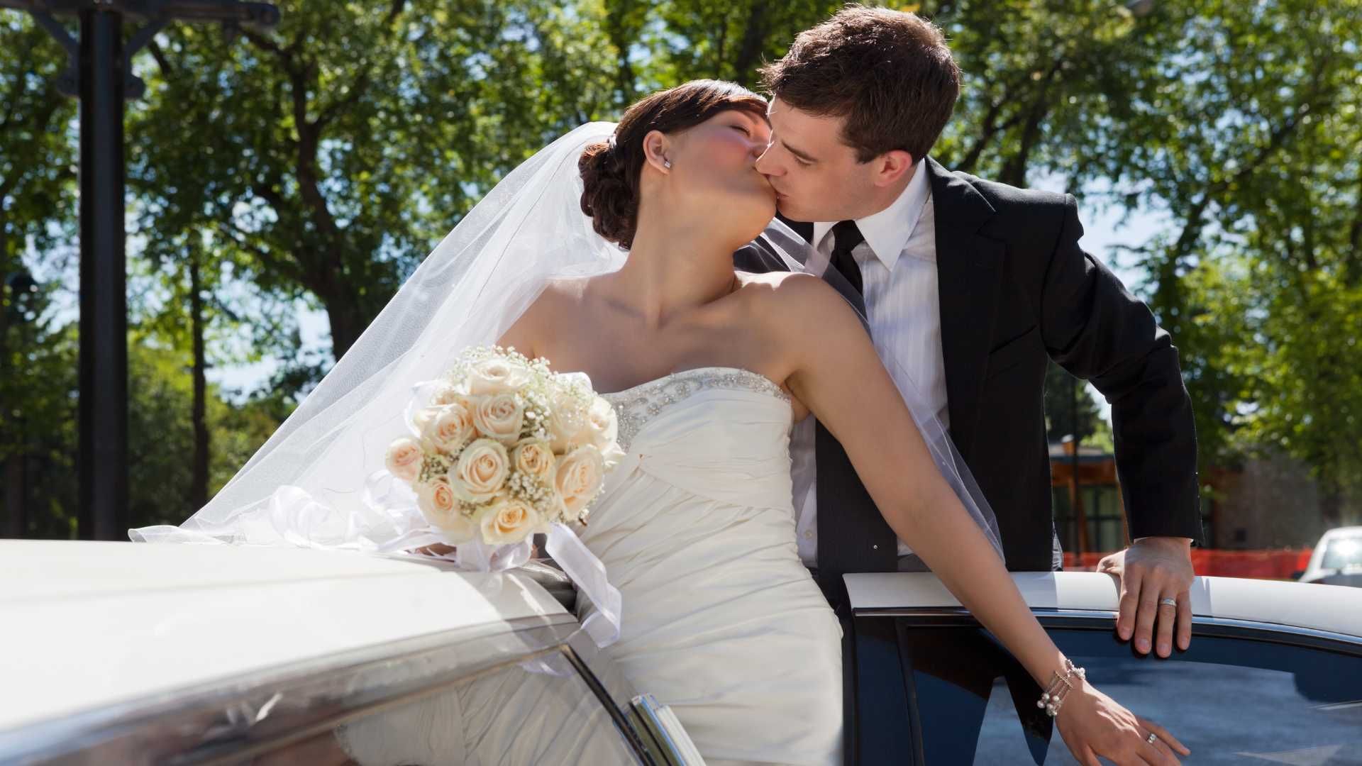 A bride and groom kissing in front of a limousine.