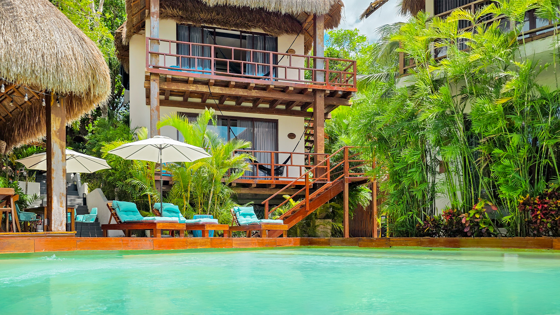 Khaban Bacalar's main pool surrounded by lush greenery, with sun loungers, umbrellas, and views of the guest rooms.