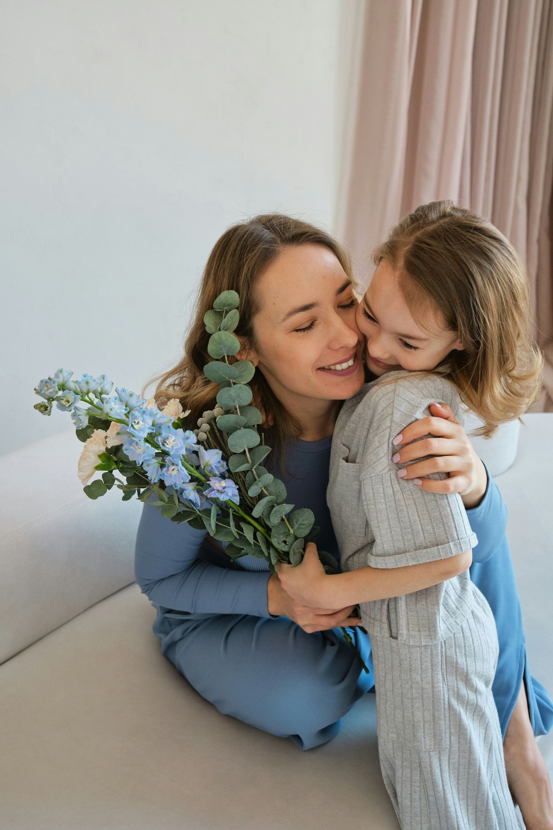 Woman hugs a girl, both smiling. Woman holds blue flowers, sitting on a bed, with beige curtains.
