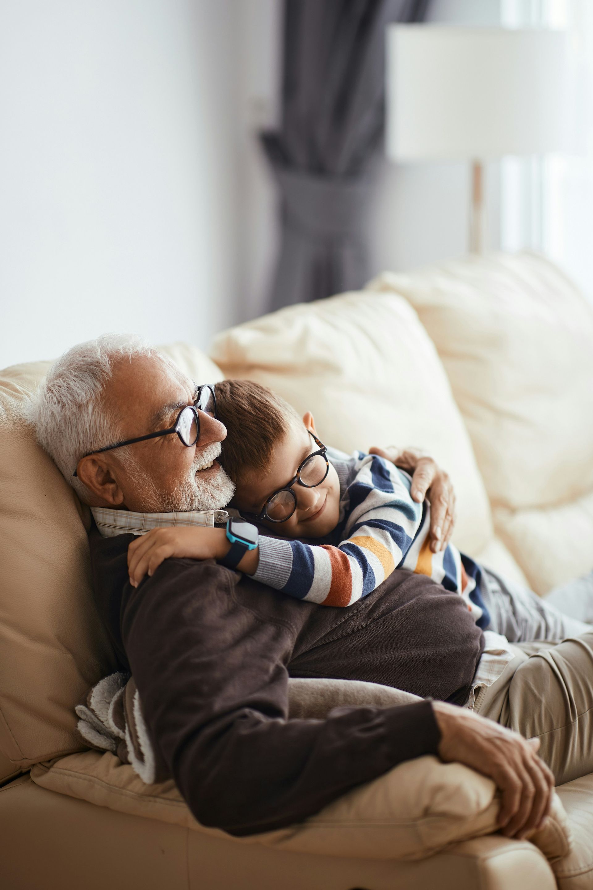 Grandfather and grandson wearing glasses embrace on a couch. The boy hugs the grandfather.