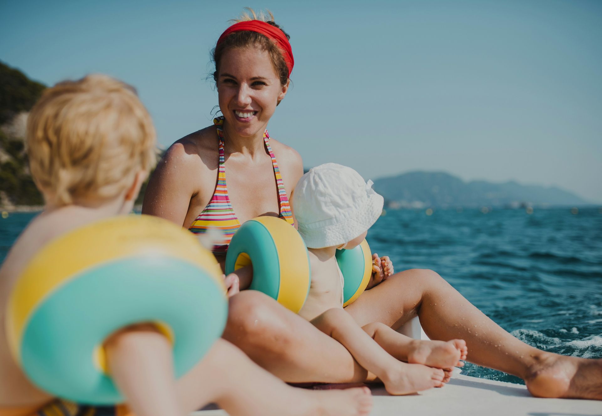 Woman and two children on a boat, smiling, wearing swimsuits, blue water, sunny day.