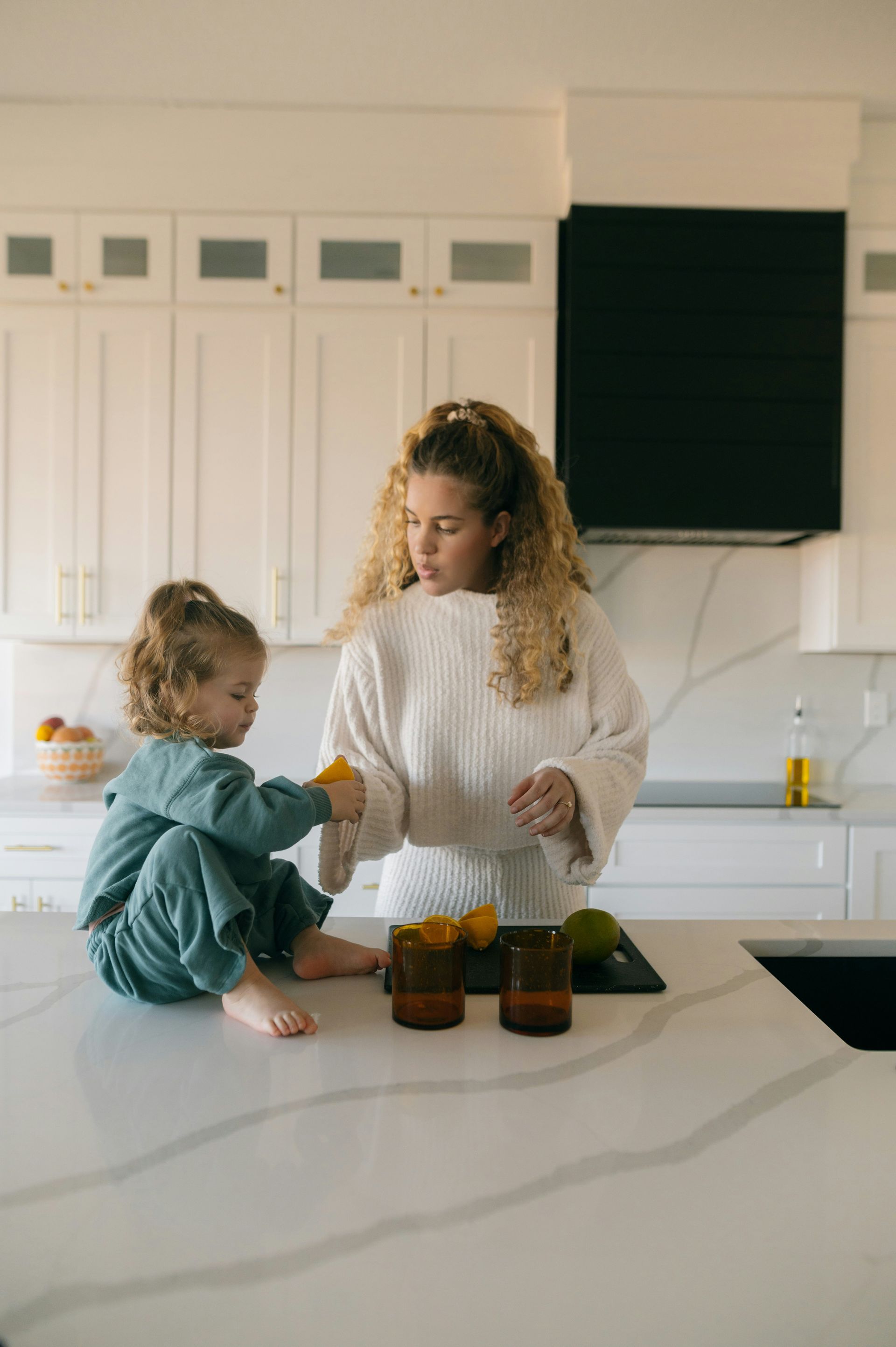 Woman and child in white kitchen, squeezing lemons. Woman has curly blonde hair. Child wears blue pajamas.